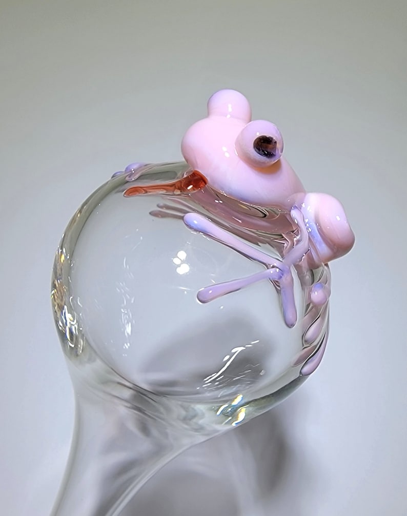 May include: A clear glass sphere topped with a sculpted light pink glass frog. The frog features smooth, rounded limbs and dark, circular eyes, resting its body and legs over the curved surface of the transparent glass orb structure.