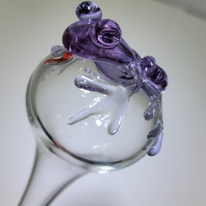 May include: A clear glass garden stake topper featuring a sculpted purple glass frog perched on top. The frog has prominent eyes and its limbs are draped over the sides of the clear glass sphere. A small red accent is visible near the frog's head.