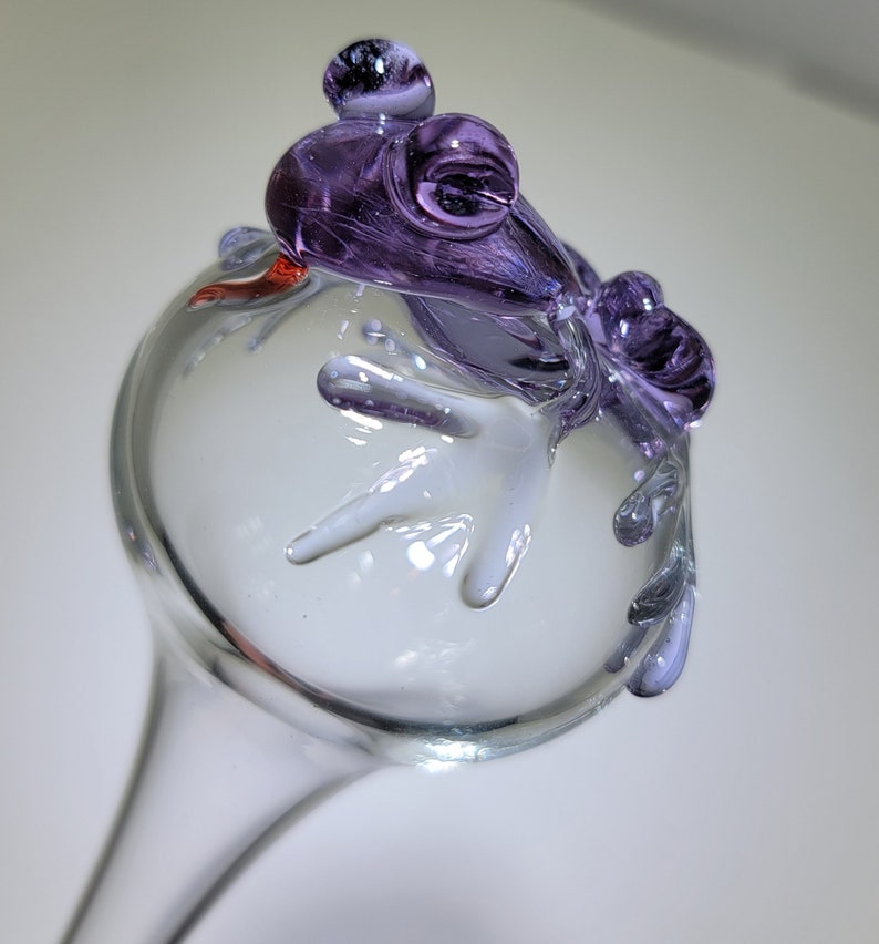 May include: A clear glass garden stake topper featuring a sculpted purple glass frog perched on top. The frog has prominent eyes and its limbs are draped over the sides of the clear glass sphere. A small red accent is visible near the frog's head.