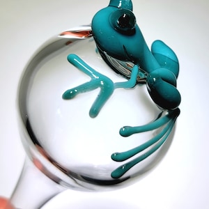 May include: A clear glass wine glass featuring a sculpted teal glass frog perched on the side of the bowl. The frog has a smooth, rounded body with elongated limbs and digits that extend across the surface of the transparent glass bowl.
