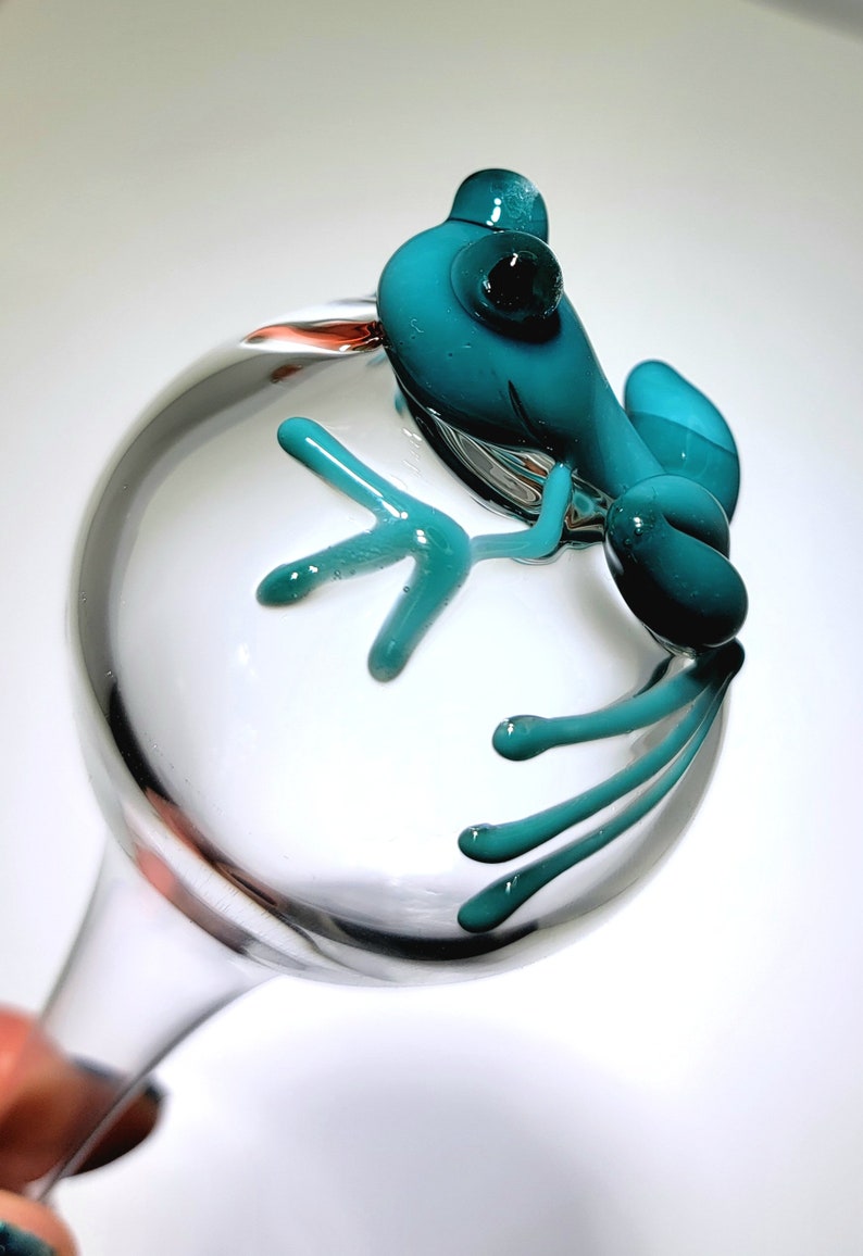 May include: A clear glass wine glass featuring a sculpted teal glass frog perched on the side of the bowl. The frog has a smooth, rounded body with elongated limbs and digits that extend across the surface of the transparent glass bowl.