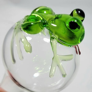 May include: A clear glass globe plant watering spike featuring a sculpted green glass frog perched on top. The frog has dark, round eyes and detailed limbs that extend over the surface of the spherical glass bulb, which is transparent.