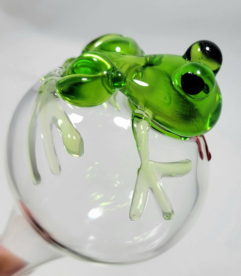 May include: A clear glass globe plant watering spike featuring a sculpted green glass frog perched on top. The frog has dark, round eyes and detailed limbs that extend over the surface of the spherical glass bulb, which is transparent.