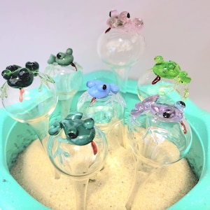 May include: A collection of seven glass plant watering globes, each topped with a small, colorful glass frog figure. The frogs are in various colors including dark green, light green, blue, purple, and pink, all featuring small red tongues.