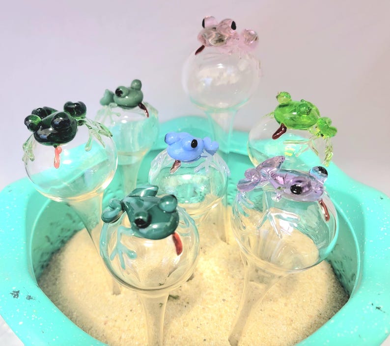 May include: A collection of seven glass plant watering globes, each topped with a small, colorful glass frog figure. The frogs are in various colors including dark green, light green, blue, purple, and pink, all featuring small red tongues.