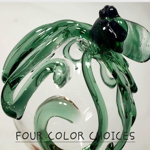 May include: A clear glass sculpture shaped like an octopus with long, curved, translucent green tentacles and a dark green head. The text "FOUR COLOR CHOICES" is printed in black at the bottom of the image, underlined by a thin black line.