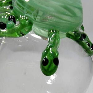May include: Close-up of a green glass turtle figurine with black eyes. The turtle is sitting on a clear glass base.