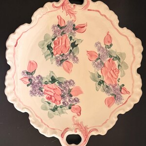 May include: An off-white ceramic serving tray with a scalloped edge and two integrated handles. The surface features hand-painted floral clusters of pink roses and purple lilac blossoms with green leaves, accented by pink scrollwork near the handles.