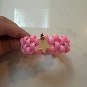 Könnte beinhalten: Ein Armband aus Reihen hellrosa Kunststoffperlen, die mit einem violetten Gummiband verflochten sind. Ein einzelner, fünfzackiger Sternanhänger mit blasser, halbtransparenter Oberfläche ist mit einem kleinen Metallring in der Mitte befestigt.