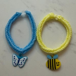 Könnte beinhalten: Zwei Gummiband-Armbänder mit Fischgrätenmuster. Ein Armband ist hellblau und hat einen weißen und blauen Schmetterlingsanhänger. Das andere Armband ist blassgelb und hat einen gelben, schwarzen und hellblauen Bienenanhänger mit einem lächelnden Gesicht.