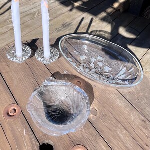 Könnte beinhalten: Zwei Kerzenhalter aus klarem Glas mit weißen Stabkerzen, eine ovale Servierplatte aus klarem Glas mit eingraviertem Blumenmuster und eine Schale aus klarem Glas mit einem spiralförmigen Rippenmuster auf der Innenfläche.