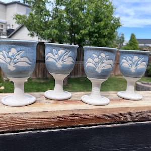 Puede incluir: Cuatro cálices de cerámica con un esmalte azul claro moteado en el cuenco y una base blanca sólida. Cada cuenco está decorado con un patrón floral en relieve de color blanco. Los cálices tienen un borde acampanado, un tallo corto y pie.