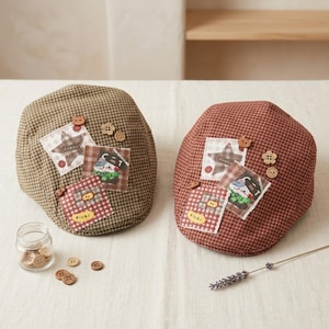 May include: Two flat caps in olive green and reddish-brown checkered fabric, each decorated with three fabric patches featuring star and cat illustrations, plus small wooden buttons. A glass jar filled with extra wooden buttons sits nearby.