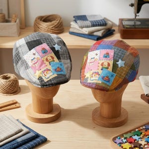 May include: Two flat caps displayed on wooden stands. The left cap is grey and black plaid with a pink patch featuring cartoon characters and star-shaped appliqués. The right cap is a multi-colored plaid with a similar pink patch and star appliqués.