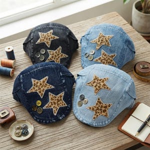 May include: Four denim flat caps in varying shades of blue and black, each featuring two leopard-print star appliqués with rhinestone borders and a cluster of three small round buttons attached to the fabric surface of the headwear.