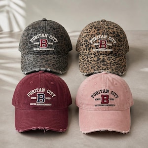 May include: Four baseball caps with distressed brims. Two feature leopard print patterns in gray and brown. Two are solid colors in maroon and dusty pink. Each cap displays embroidered text reading PURITAN CITY SINCE 1630 BOSTON MASSACHUSETTS.