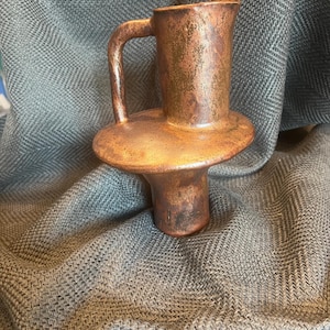 May include: A copper-toned ceramic pitcher with a unique structural design featuring a wide, flat circular disc separating the upper cylindrical body from the lower base, complete with a curved handle attached to the side of the upper section.