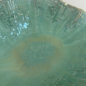 May include: A round ceramic bowl with a glossy turquoise glaze and a textured, geometric relief pattern across the interior surface. The center of the bowl features a subtle, lighter beige-toned wash that blends into the vibrant blue-green finish.