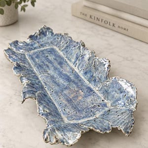 May include: A rectangular ceramic tray with a mottled blue and white glaze. The tray features a textured, uneven rim that mimics the appearance of ruffled leaves, with a flat, speckled base and a glossy finish throughout the entire surface.