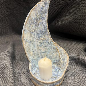 Può includere: Un portacandele in ceramica a forma di mezzaluna con una finitura smaltata maculata blu, bianca e beige. Il pannello posteriore curvo si eleva verticalmente da una base circolare che contiene una piccola candela cilindrica bianca con stoppino.