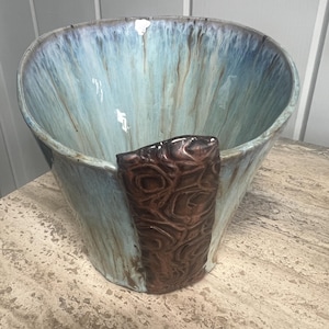 May include: A ceramic bowl with a wide, flared rim featuring a glossy light blue and white drip glaze finish. A prominent, textured dark brown vertical accent piece with a swirling, organic relief pattern is attached to the front exterior.