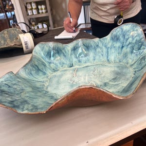 May include: A shallow ceramic bowl with a wavy, irregular rim. The interior features a mottled light blue and white glaze with a stamped dandelion design at the centre. The exterior has a textured, metallic copper-toned finish.
