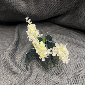 Può includere: Un vaso in ceramica blu scuro con una forma ondulata e organica, caratterizzato da una superficie strutturata con piccole rientranze. Il vaso contiene un rametto di fiori artificiali bianchi con molteplici piccoli petali.