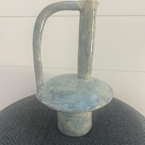 May include: A light blue ceramic vase with a speckled finish, featuring a wide circular base, a narrow cylindrical neck, and a tall, arched handle that connects the top of the neck to the side of the base, creating a unique, asymmetrical silhouette.