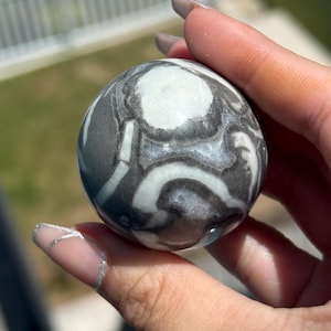 May include: A polished spherical stone featuring a high-contrast pattern of dark charcoal gray and creamy white swirling bands. The smooth, rounded surface displays organic, flowing shapes that wrap around the entire circumference of the orb.