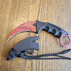 May include: A curved karambit knife featuring a red blade with a black spiderweb pattern and a black textured handle with a finger ring. The knife is accompanied by a black sheath with a black cord attached for carrying or securing the item.