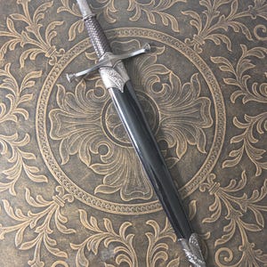 May include: A dagger with a textured silver-toned grip, a crossguard, and a pommel. The blade is housed in a smooth black scabbard featuring ornate silver-toned metal fittings at the throat and the chape, displaying intricate embossed scrollwork.