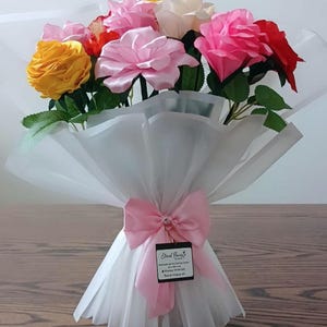 May include: A bouquet of artificial roses in yellow, pink, white, and red, featuring green fabric leaves. The flowers are wrapped in translucent white paper, tied with a pink satin ribbon bow, and include a small square tag labelled Eternal Flowers.