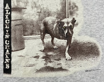 Può includere: T-shirt grigio con una grafica in bianco e nero di un cane con gli occhi gialli. Il testo "ALICE IN CHAINS" è stampato verticalmente a sinistra. L'immagine ha un effetto invecchiato, che conferisce alla t-shirt un aspetto vintage.