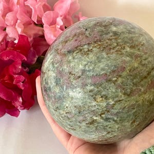 May include: A polished spherical stone featuring a mottled texture with shades of sage green, olive, and deep magenta. The surface displays natural mineral inclusions and crystalline patterns throughout the round form, showcasing a smooth, reflective finish.
