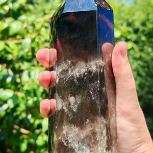 May include: A tall, hexagonal smoky quartz crystal point with a dark, translucent brown colour. The crystal features a polished, pointed top and a natural, textured base with visible internal inclusions and cloudy white mineral formations.