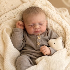 Puede incluir: Una muñeca recién nacida dormida con piel clara y cabello fino, que viste un mameluco gris de manga larga estilo henley con tres pequeños botones de madera en el cuello. La muñeca está envuelta en una manta texturizada de color crema y sostiene un pequeño juguete de tela color crema con forma de conejo con rasgos cosidos simples.