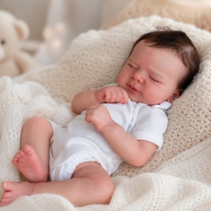 Peut inclure: Une poupée nouveau-né réaliste aux cheveux foncés et aux yeux fermés, vêtue d'un body blanc uni à manches courtes. La poupée est allongée dans une position inclinée, reposant sur une couverture tricotée crème au motif texturé.