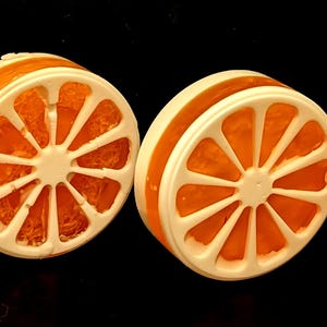 Può includere: Quattro saponette circolari a forma di spicchi d'arancia, caratterizzate da una cornice bianca con un design segmentato e un centro arancione brillante e traslucido che imita l'aspetto della polpa degli agrumi su uno sfondo nero.