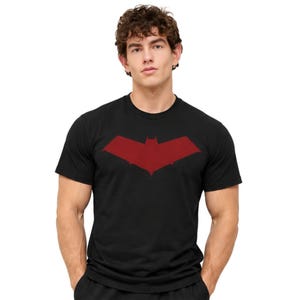May include: A black short-sleeved t-shirt featuring a dark red graphic of a stylised bat-like emblem centred on the chest. The garment has a classic crew neckline and a straight hem, constructed from a solid black fabric with a matte finish.