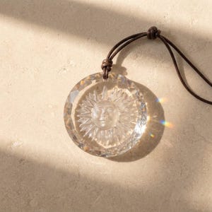 May include: A clear, circular glass pendant featuring a relief carving of a sun with a face in the center. The pendant has faceted edges and is suspended from a thin, dark brown cord that is knotted at the top of the circular frame.