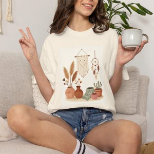 May include: An off-white short-sleeve t-shirt featuring a graphic print of a macrame wall hanging, a dreamcatcher, and several terracotta-colored vases containing pampas grass, dried flowers, and a small green succulent plant.