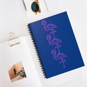 May include: A spiral-bound notebook with a dark blue cover featuring three stacked pink neon-style flamingo outlines. The notebook is open, revealing white pages, one of which displays a small square photograph of a desert building.