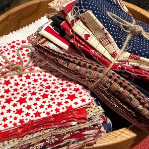 May include: Two stacks of folded fabric squares tied with twine. One stack features a red star pattern on a white background. The other stack includes various patterns in red, white, and blue, such as polka dots, stripes, and small heart motifs.