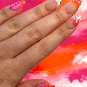 Peut inclure: Ongles en amande arborant divers motifs de nail art. Le pouce et l'auriculaire présentent un dégradé rose et orange avec des taches. L'index et le majeur affichent une french manucure avec des rayures zébrées noires, tandis que l'annulaire présente une french manucure avec une petite fleur orange.