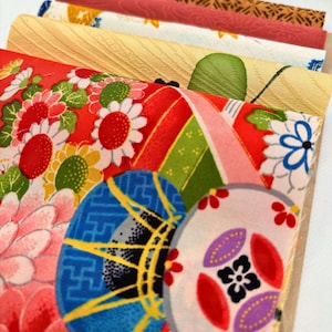 May include: A stack of folded fabric pieces featuring various patterns. The top layer displays a vibrant red background with colorful floral motifs, blue circular designs, and green accents. Beneath it are layers of yellow, white, and red textured fabrics.