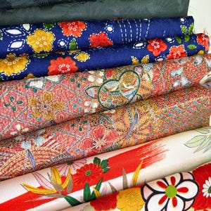 May include: A stack of folded fabric bolts featuring diverse patterns. The top is dark grey, followed by dark green, a blue floral print, a red patterned textile with gold accents, a cream fabric with red brushstrokes, and a floral print with large blossoms.