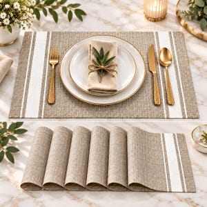 May include: A rectangular placemat featuring a woven tan texture with two vertical white stripes on each side. The placemat is shown in a table setting with a white plate, gold-toned hammered cutlery, and a folded tan napkin tied with twine.