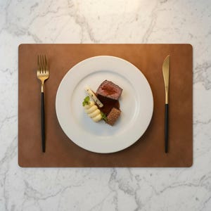 May include: A rectangular brown leather placemat featuring a white ceramic plate centered with a portion of seared meat, a piped potato garnish, and a brown sauce. A gold-toned fork and knife with matte black handles are placed on the sides.