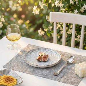 Puede incluir: Un plato redondo de cerámica con un patrón de líneas radiales en blanco y negro contiene higos frescos cortados. Una cuchara de plata, una copa de vino blanco, un gofre en un plato a juego y una vela de burbujas blanca sobre un mantel individual.