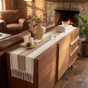 May include: A long, rectangular table runner featuring a woven cream and tan striped pattern with decorative fringe on the ends. The runner is placed on a wooden sideboard and is topped with a beige ceramic vase, two white pillar candles, and a glass.